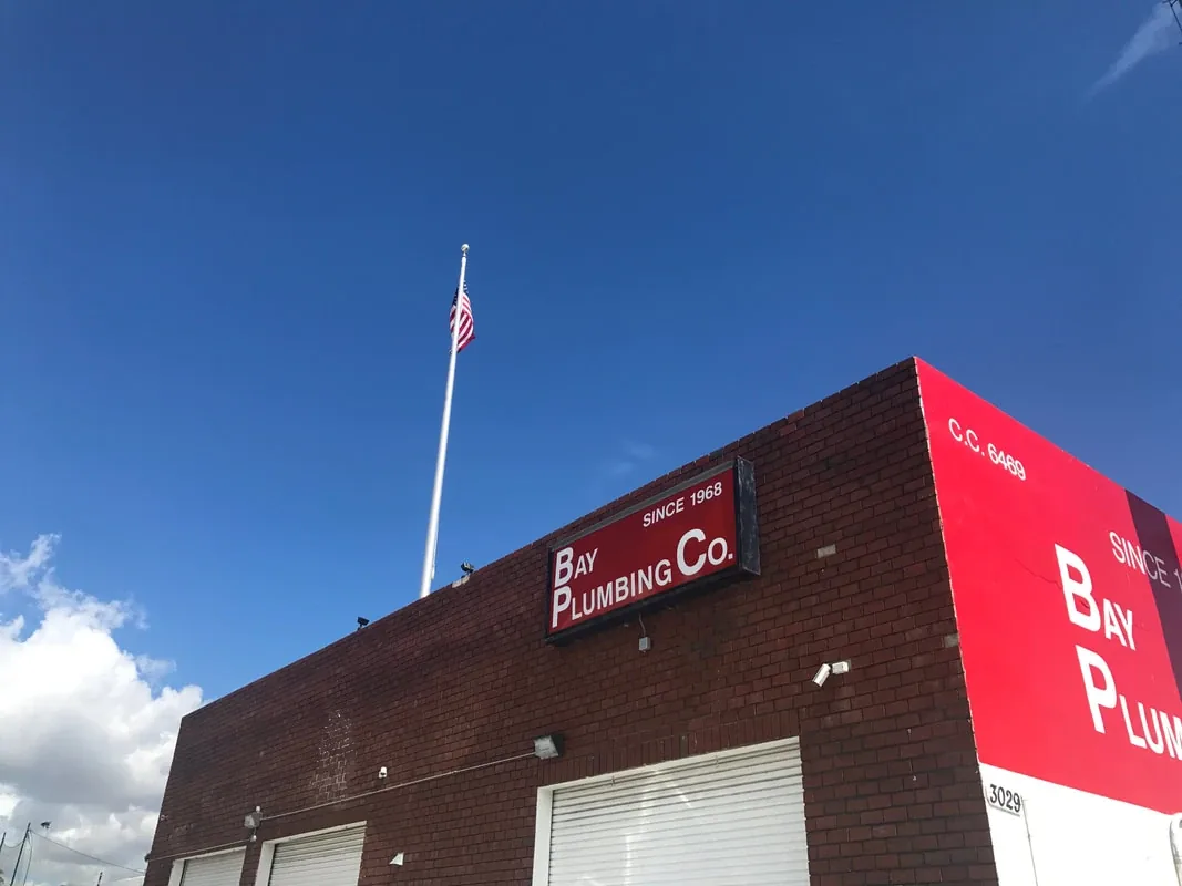 The Bay Plumbing Co. shop: a red brick building with a 'Bay Plumbing Co. Since 1968' sign, an American flag on the roof, and a red wall painted with license number C.C. 6469.